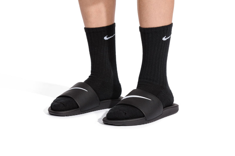 Kids black slide sandals on feet with black socks, white Nike swoosh logos