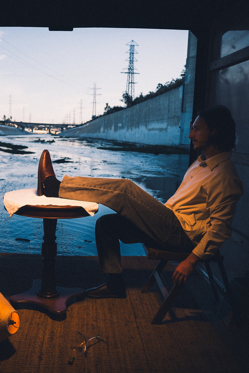 Signature mule in oxblood leather with a Vibram Mini Ripple sole worn by a man relaxing beside a river.