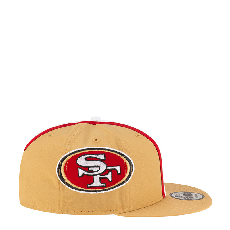 Tan baseball cap with oval team logo patch and red trim along sides