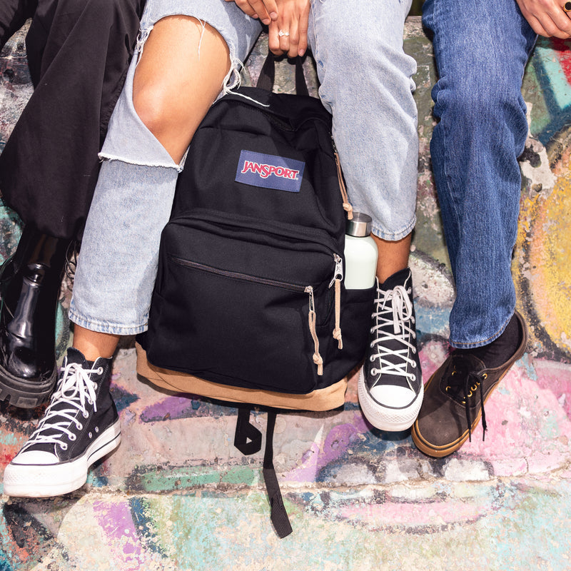 Black JanSport backpack on street with front zippers, logo patch, and side bottle