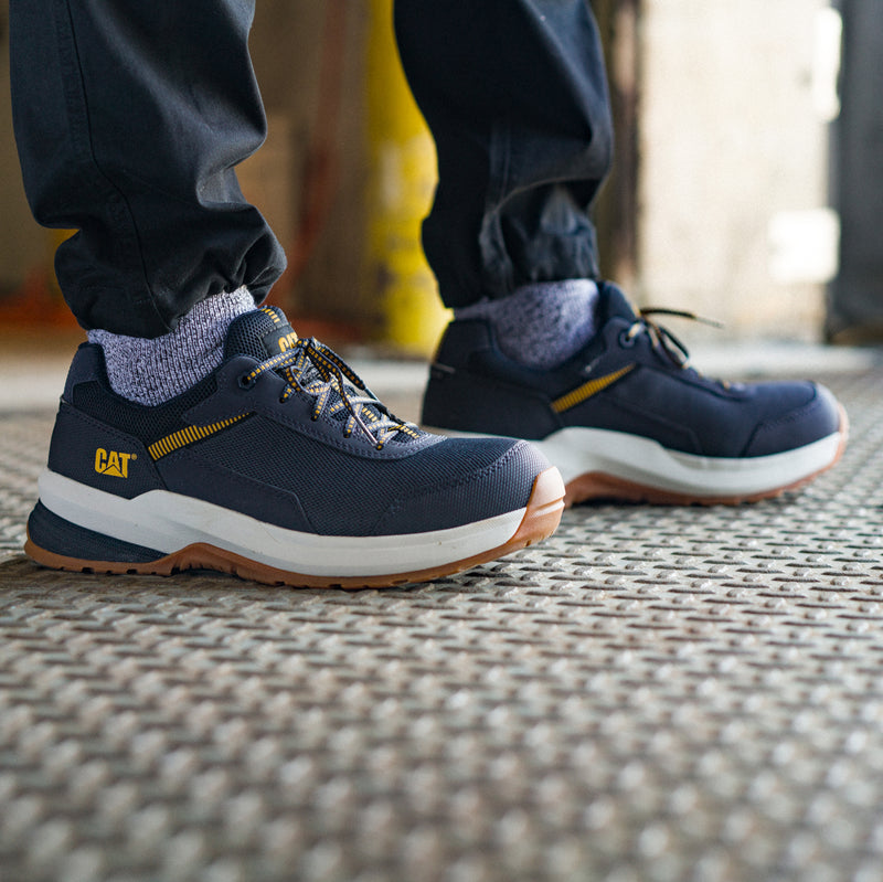 Men's CAT safety sneaker on pavement, navy upper with yellow laces, white sole