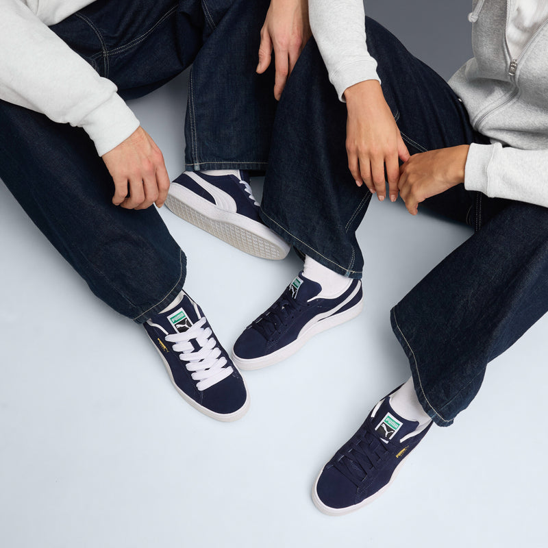 Two navy suede low-top sneakers with white accents, lace-up, worn with jeans